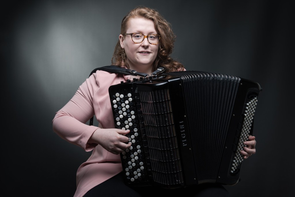 Accordionist Aine McLoughlin holding a large accordion in a pink jacket.