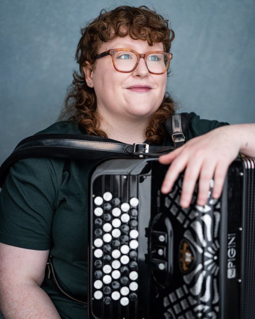 London accordion player Aine McLoughlin holding large accordion in front of blue background